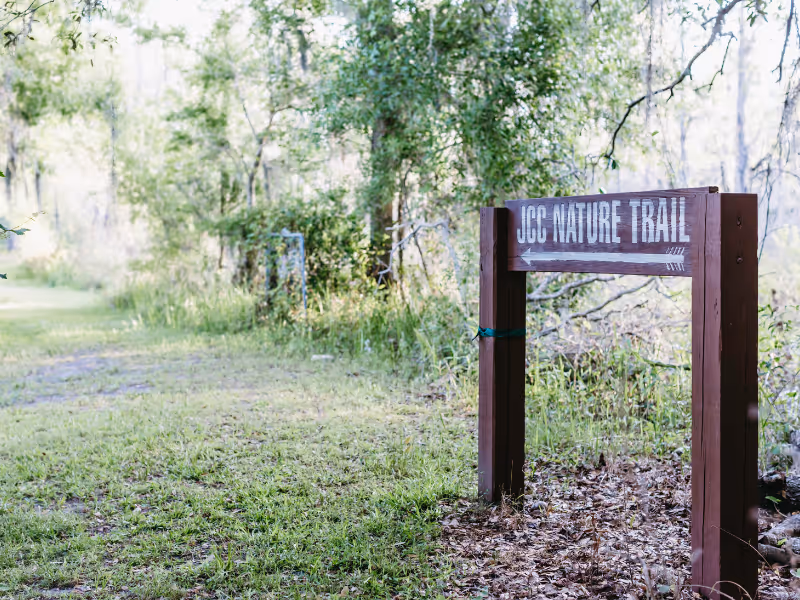 A wooden signpost with the text 'JCC NATURE TRAIL' pointing to a grassy path surrounded by trees and greenery in a natural outdoor setting.