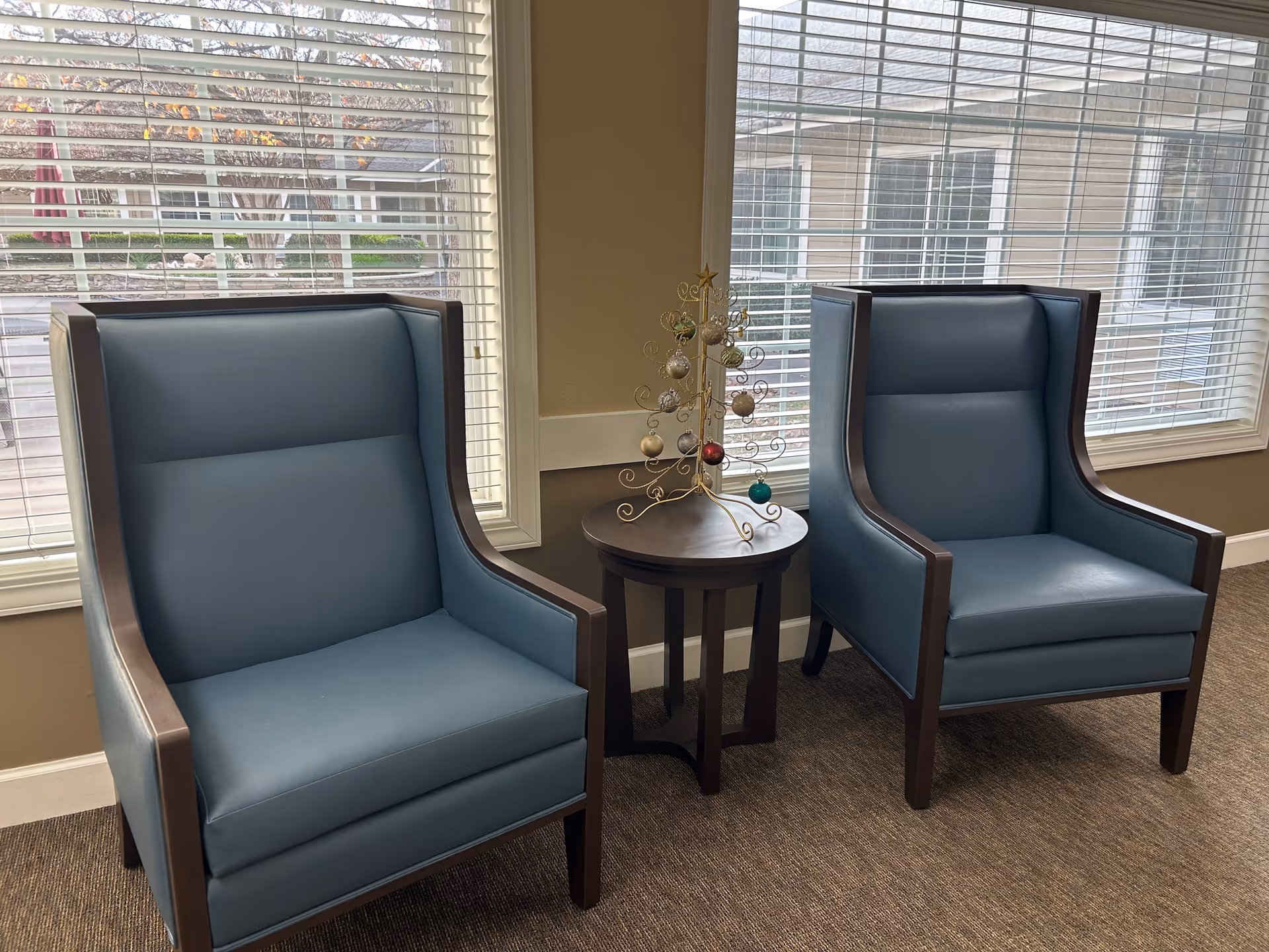 Two blue upholstered armchairs with wooden frames are positioned on either side of a small round wooden table. On the table is a decorative metal Christmas tree with colorful ornaments. Behind the chairs are large windows with white blinds, showing an outdoor view of a building and some greenery.