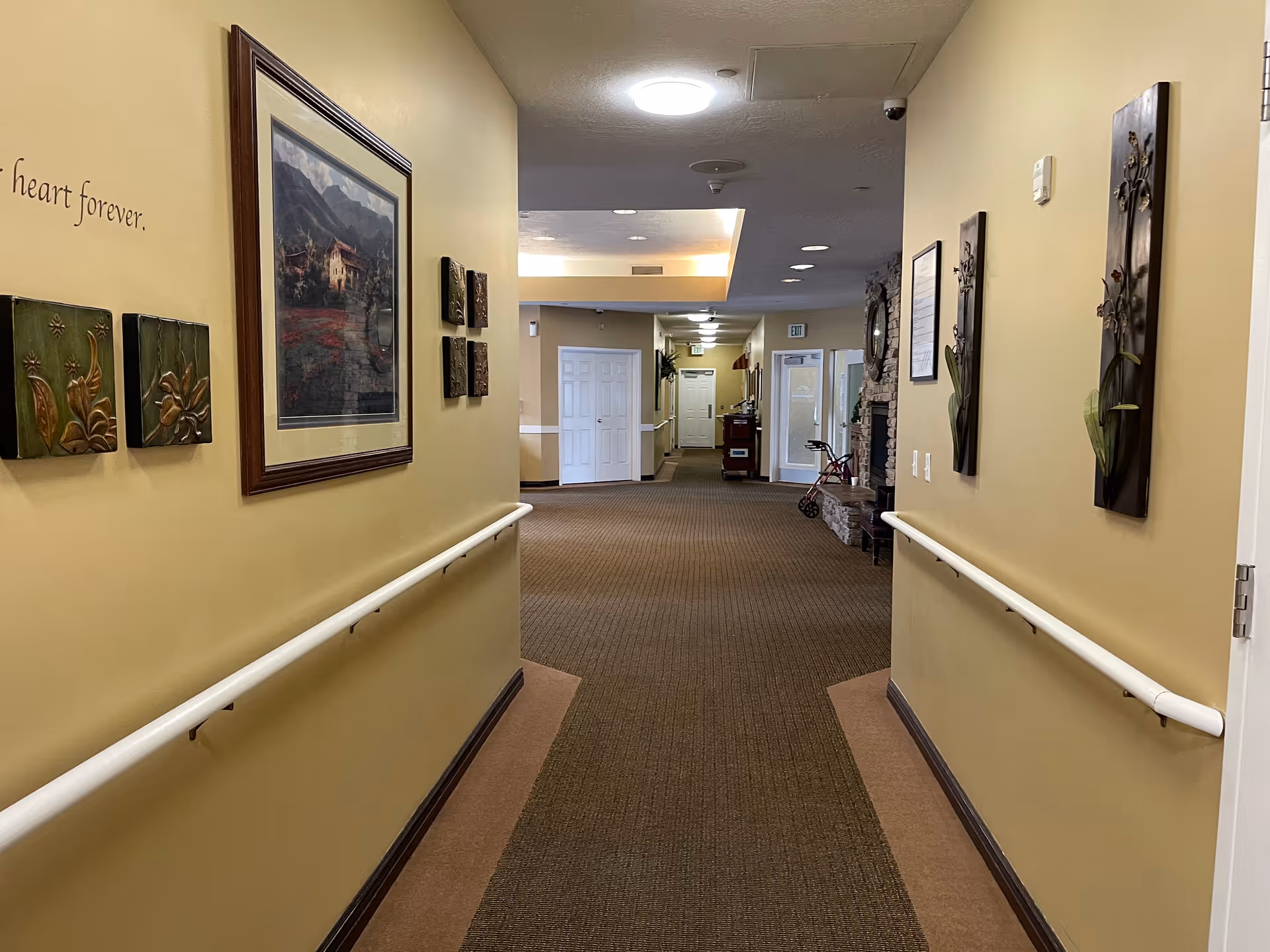 A well-lit hallway in a senior living facility with beige walls and carpeted floor. The walls are decorated with framed artwork and decorative panels. Handrails run along both sides of the hallway. At the end of the hallway, there are several doors and a walker is visible near a stone fireplace on the right side.