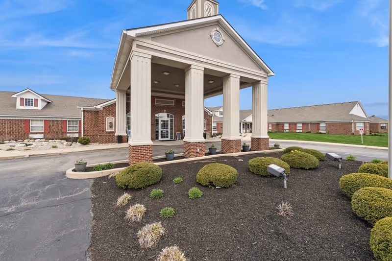 Front exterior view of Five Star Residences of Lafayette featuring a covered entrance with four large columns, brick walls, manicured bushes, and a clear blue sky.