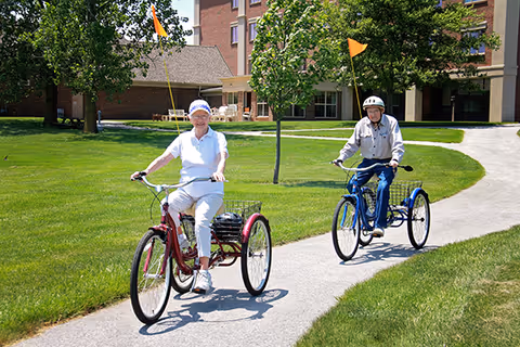 Two elderly individuals riding three-wheeled bicycles on a paved path in a grassy outdoor area with trees and a multi-story building in the background. Both riders are wearing helmets and casual clothing.
