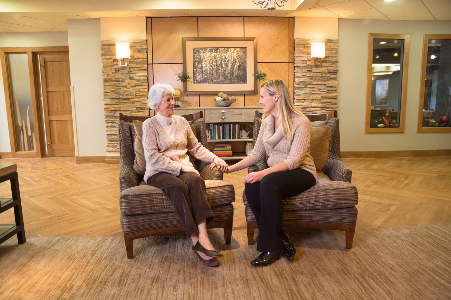 An elderly woman and a younger woman sit in a cozy lounge area holding hands and conversing.