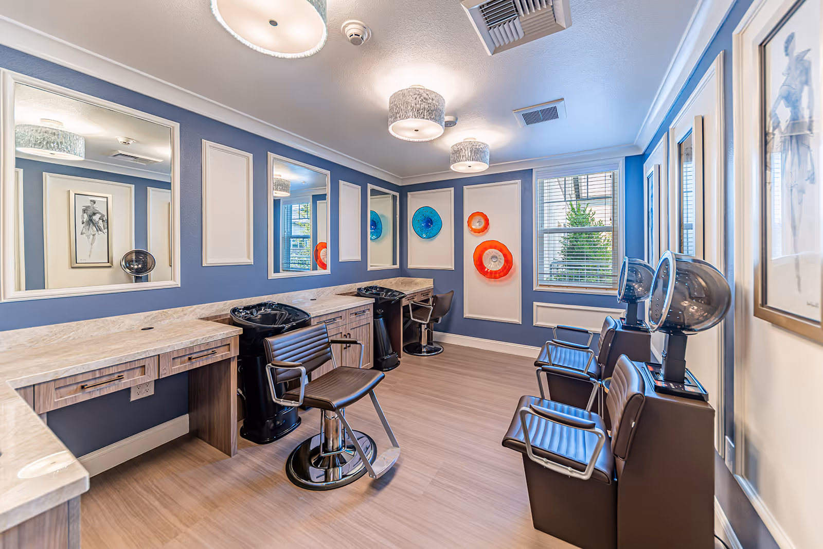Interior view of a senior living facility's hair salon with blue walls, three styling chairs, two hair washing stations, two hair dryers, large mirrors, decorative wall art, and a window letting in natural light.