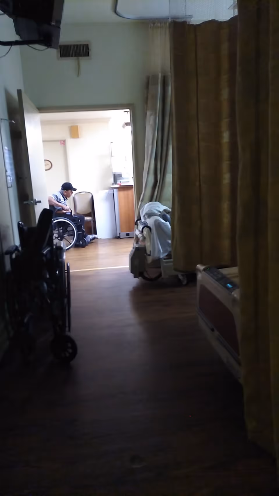 Interior view of a care facility corridor with privacy curtains, a bed and equipment, and a person sitting in a wheelchair near a doorway.