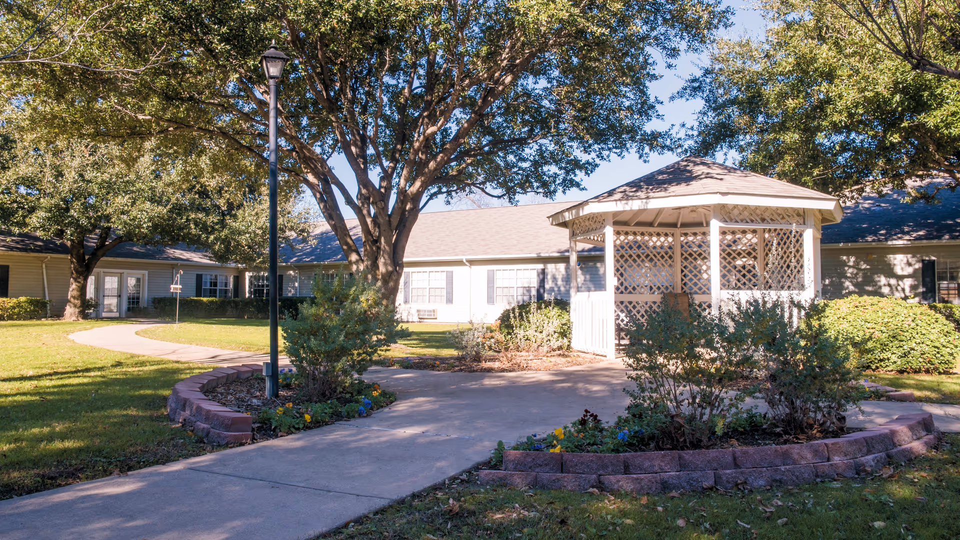 Outdoor view of a senior living facility with a paved walkway leading to a white gazebo surrounded by landscaped flower beds and bushes. Large trees provide shade, and the building with multiple windows and doors is visible in the background under a clear blue sky.