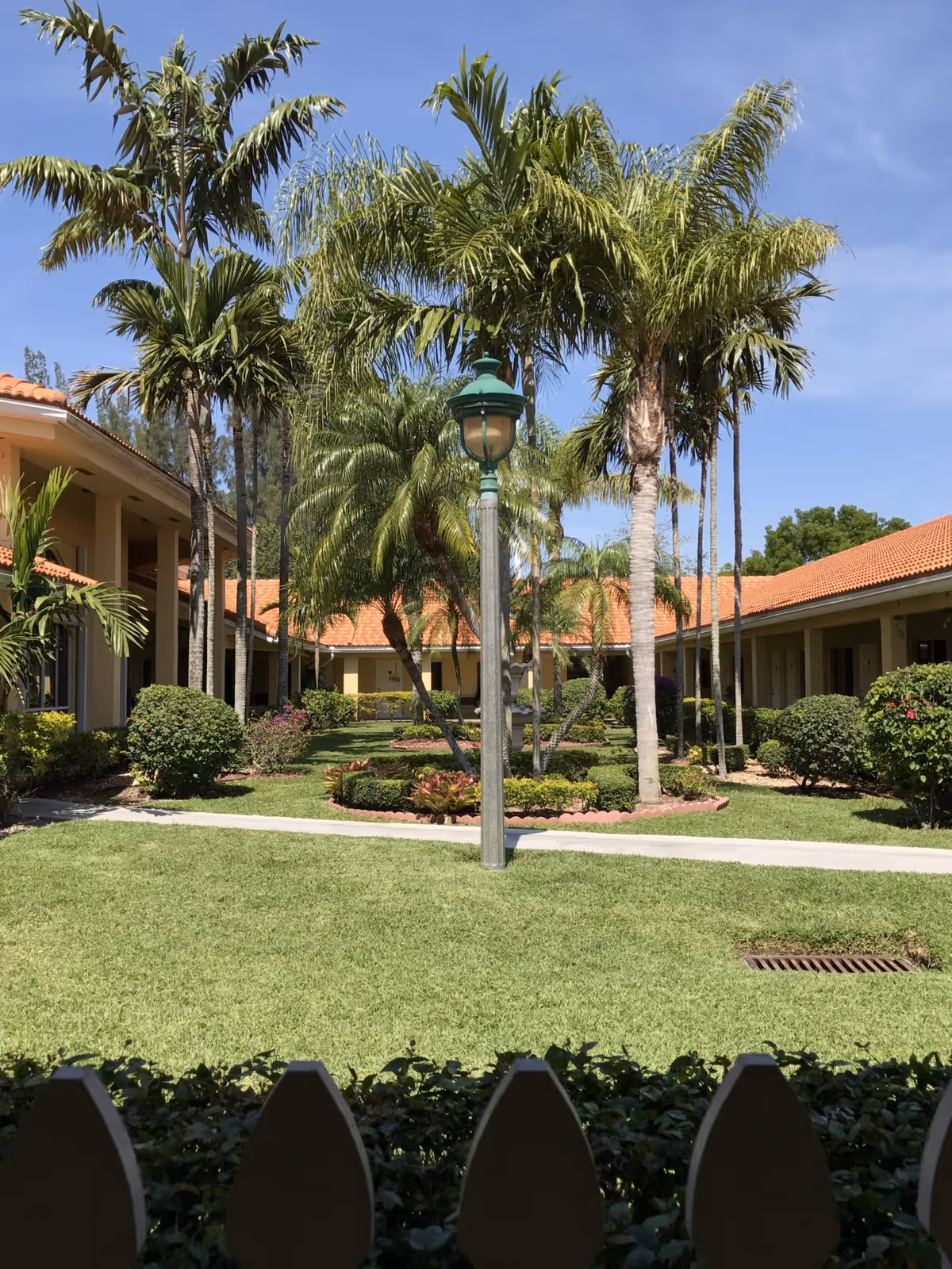 A sunny courtyard with well-maintained green grass, palm trees, and bushes surrounded by single-story buildings with orange tiled roofs. A vintage-style lamp post stands in the center of the courtyard, and a white picket fence is visible in the foreground.