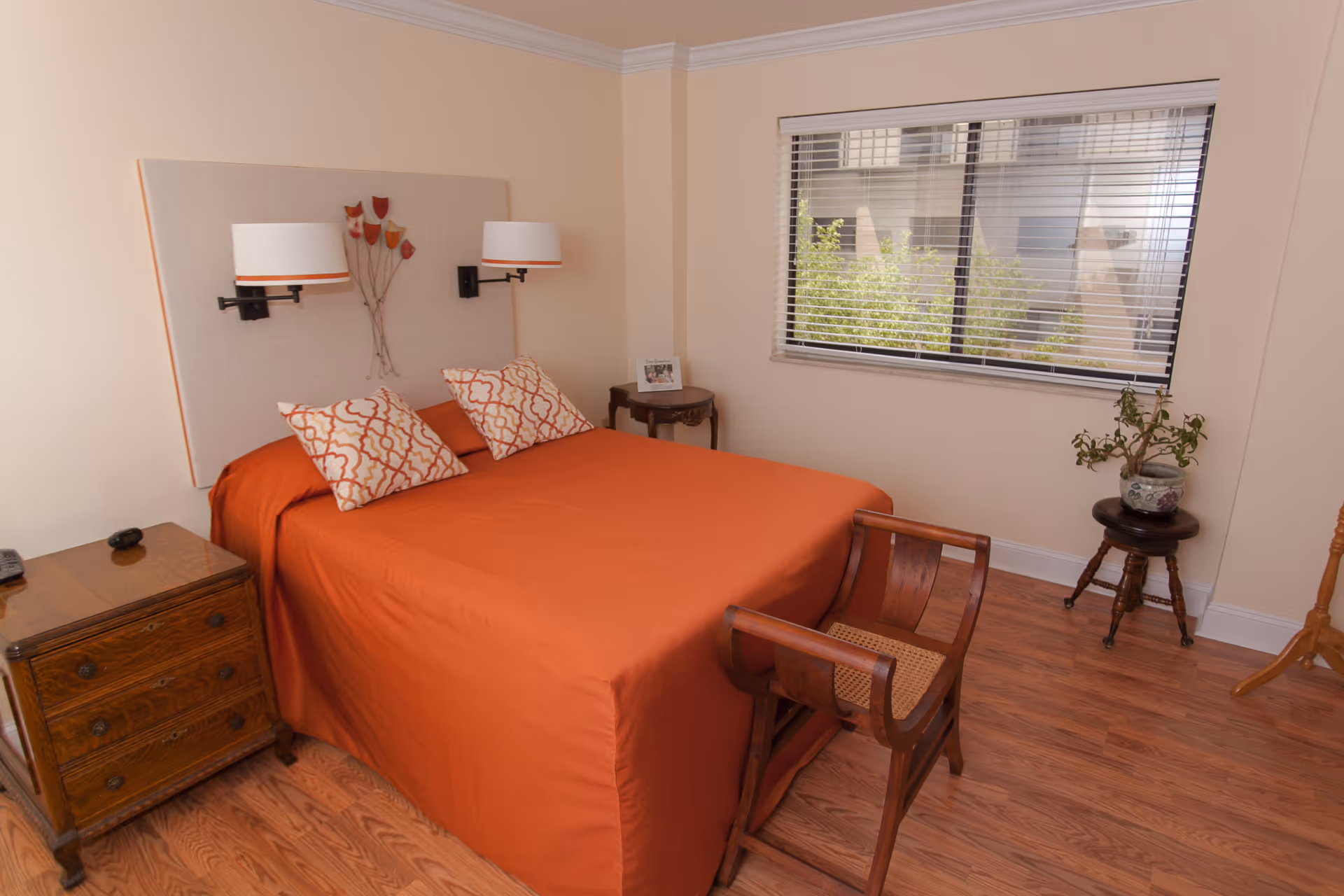 Bedroom with an orange bedspread, patterned pillows, wooden furniture, and a window with blinds.
