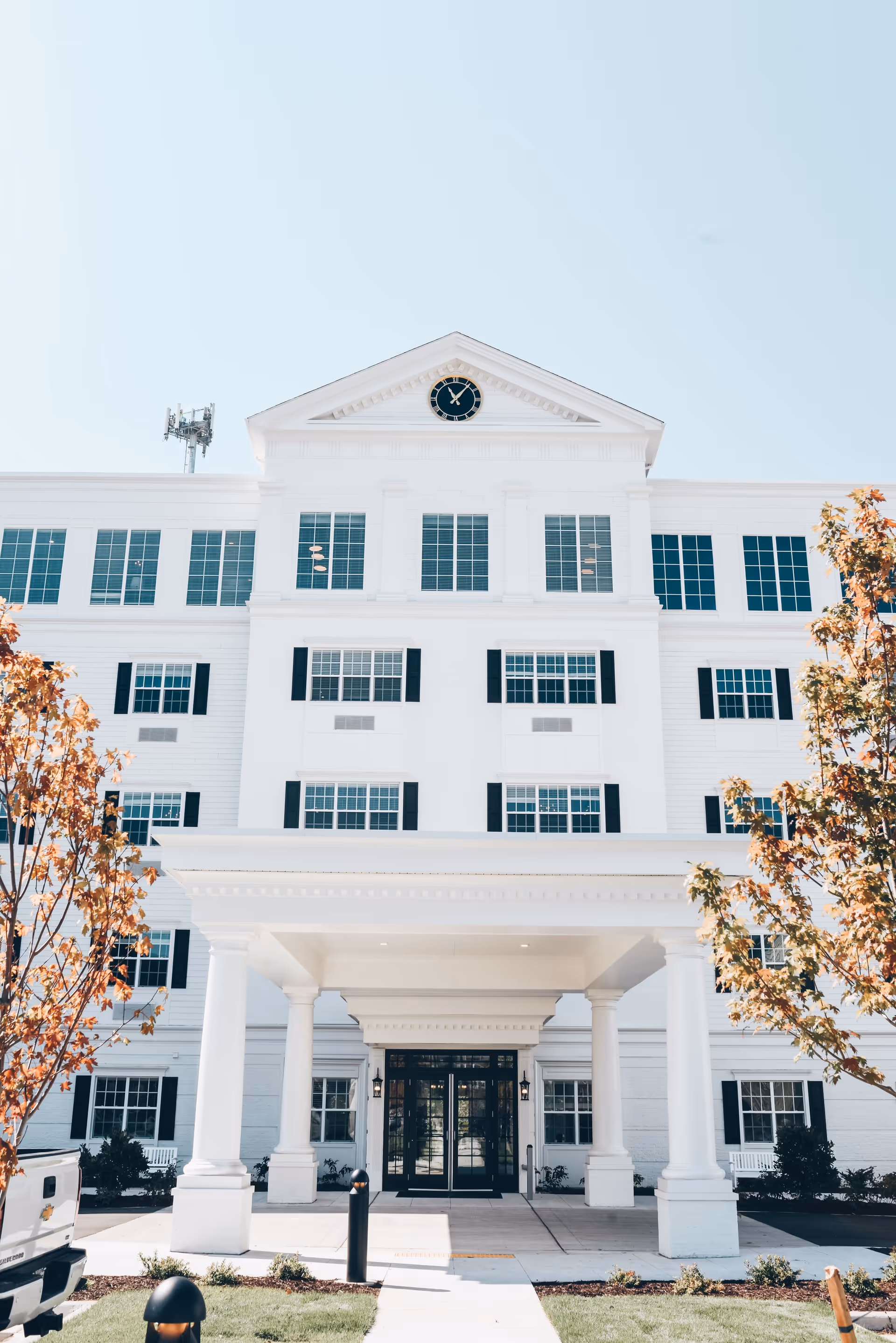 Front exterior view of a large white building with multiple windows and black shutters, a clock at the top center, a covered entrance supported by four white columns, and autumn trees on either side.
