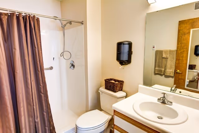 A clean bathroom featuring a white toilet with a brown woven basket on top, a white sink with a silver faucet set in a countertop with wooden trim, a large mirror above the sink, a mounted black paper towel dispenser on the wall, and a shower area with a brown shower curtain and a silver showerhead.