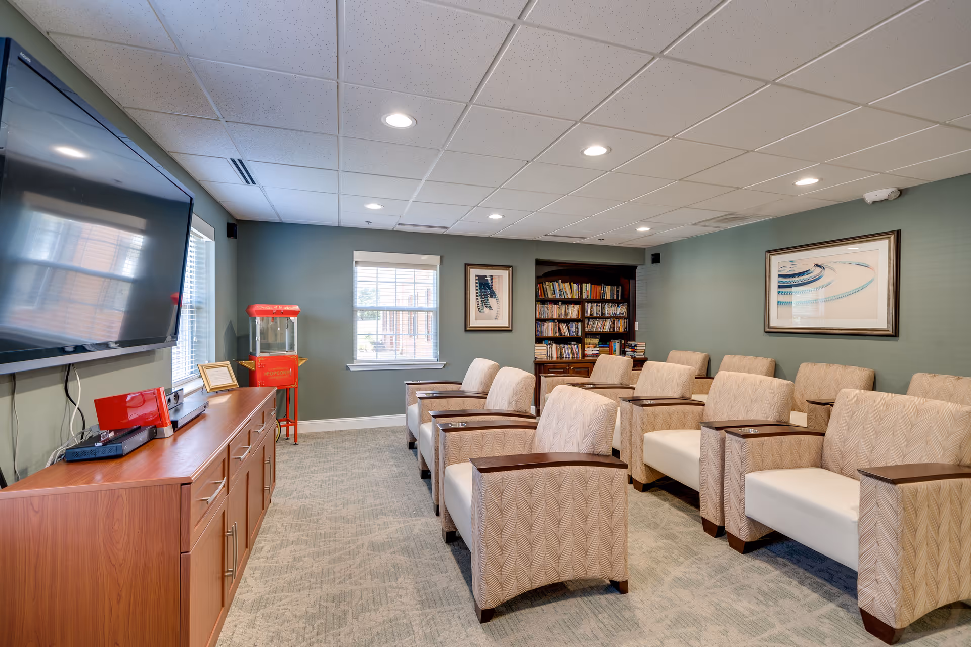 A cozy common room with multiple beige armchairs arranged in rows facing a large flat-screen TV mounted on the wall. There is a wooden cabinet beneath the TV with electronic devices on top. A red popcorn machine stands near a window with blinds. A bookshelf filled with books and framed artwork on the walls complete the room.