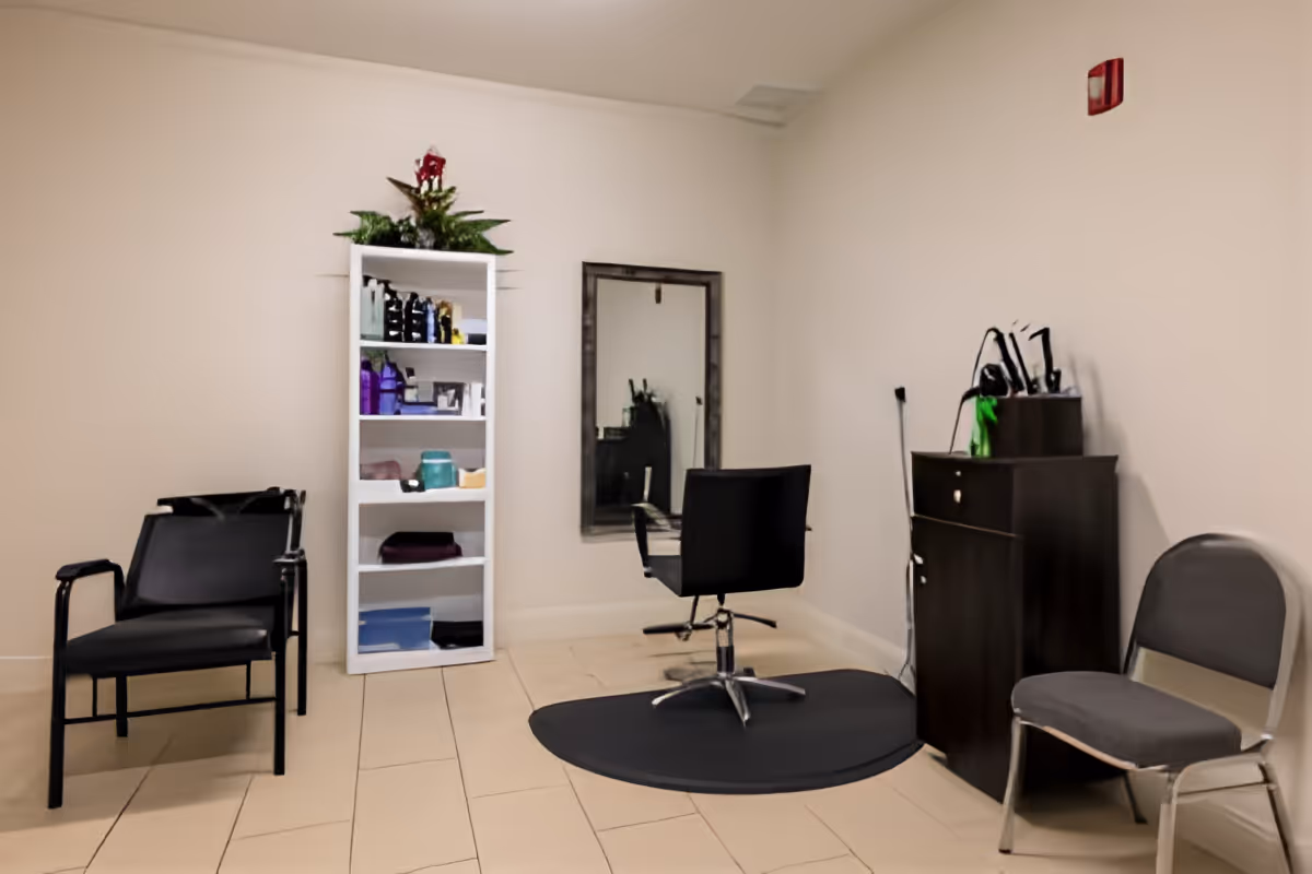 A small salon area in a facility with a black salon chair on a black mat facing a wall-mounted mirror. To the left, there are two black chairs stacked, and to the right, a gray chair and a dark wooden cabinet with hair styling tools on top. A white shelving unit with various hair products and a small plant on top is against the wall.