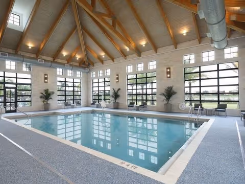 Indoor swimming pool in a bright room with a vaulted wood-beamed ceiling, large paneled windows, and seating along the sides.