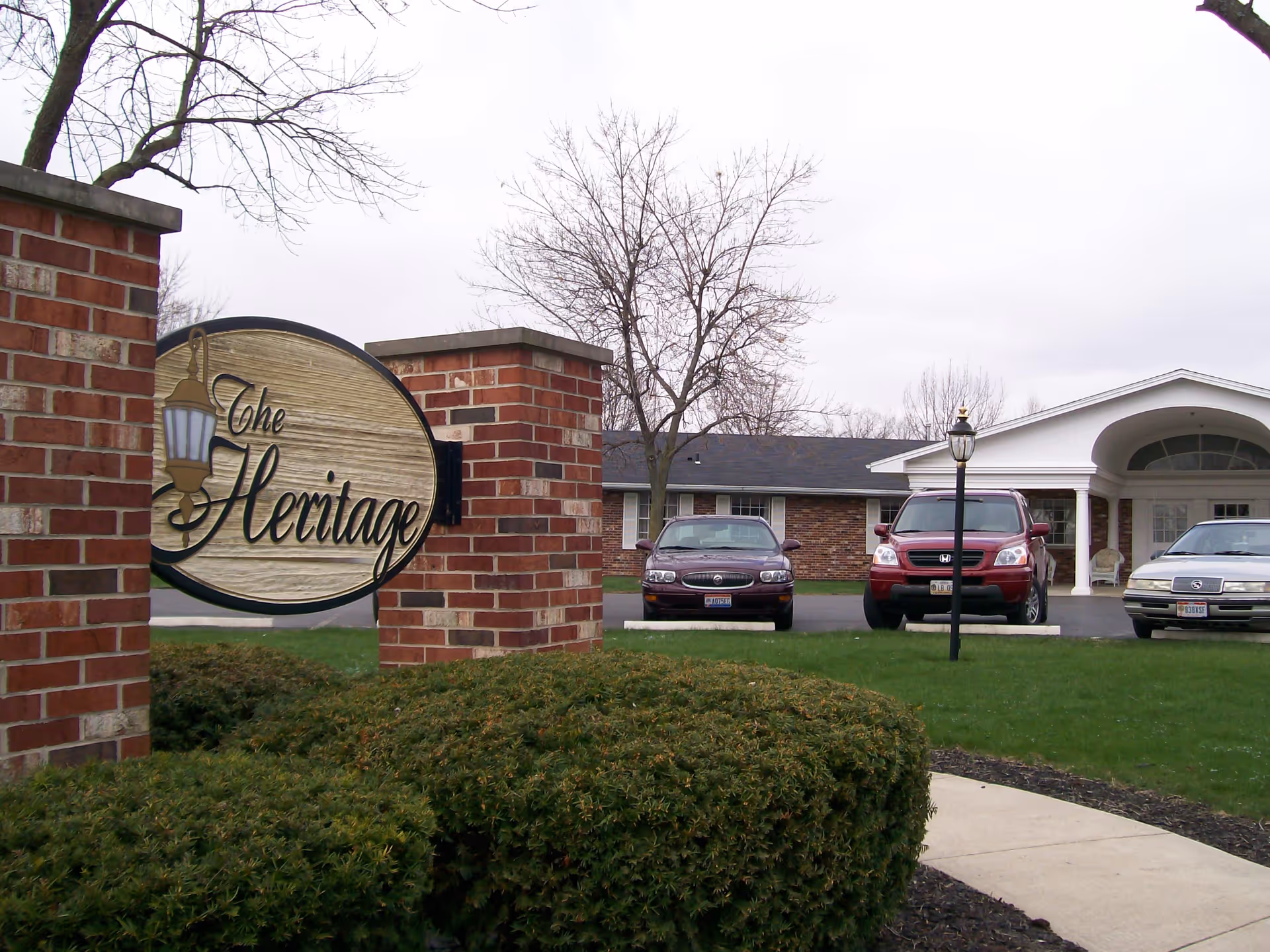 Exterior view of The Heritage facility showing a brick sign with the facility name, trimmed bushes, a sidewalk, a parking area with three cars, and a building entrance with white columns and a covered porch.