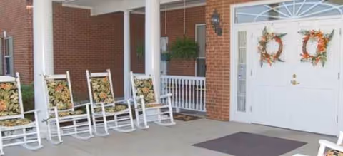 Front porch of a brick senior living facility with white rocking chairs and wreaths.