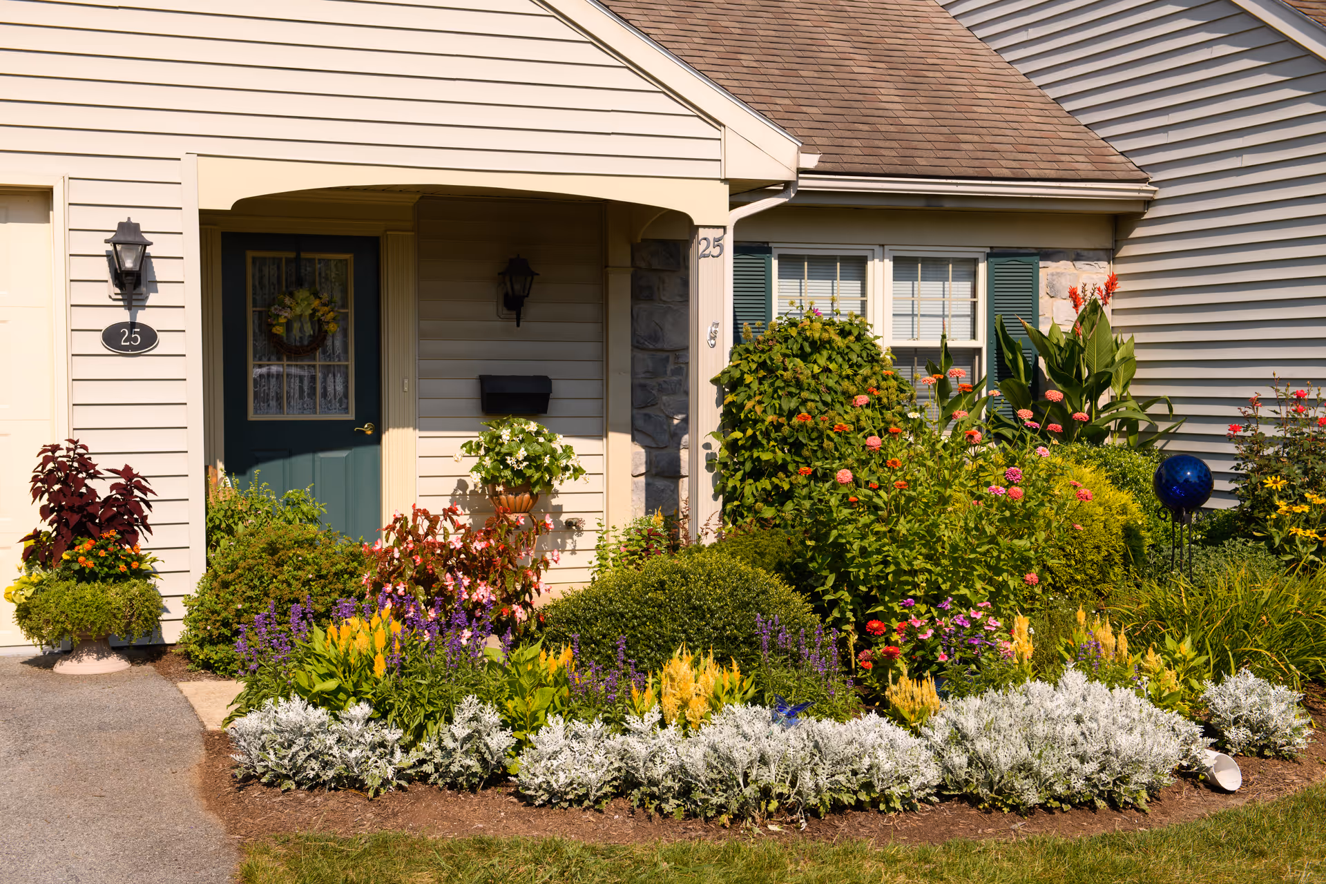 Front entrance of a residential unit at Garden Spot Village with a green door adorned with a wreath, surrounded by beige siding and a variety of colorful flowering plants and shrubs in a well-maintained garden bed.