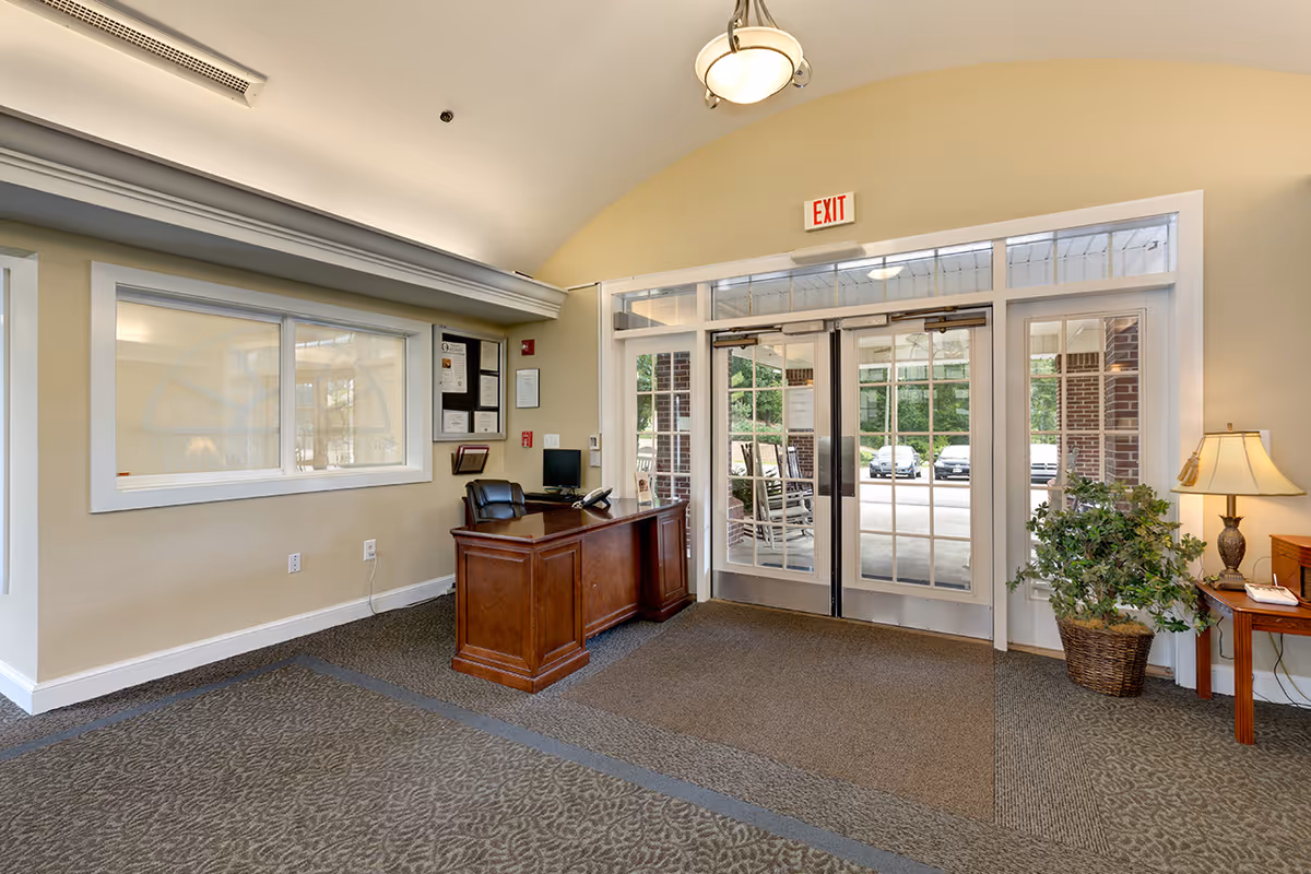 Interior view of a facility entrance area with a wooden reception desk and chair on the left, large glass double doors with an exit sign above, a potted plant and a side table with a lamp on the right, and a carpeted floor.