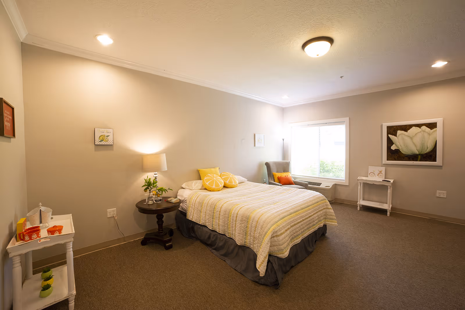 A cozy senior living bedroom with a bed covered in a striped yellow and white quilt, two lemon-shaped pillows, a round wooden side table with a lamp and plant, a gray armchair with colorful cushions near a window, and simple wall decorations including framed pictures and a flower painting.