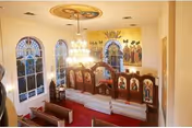 Interior view of a chapel with an iconostasis, stained-glass windows, mural, a central chandelier, and wooden pews on red carpet.