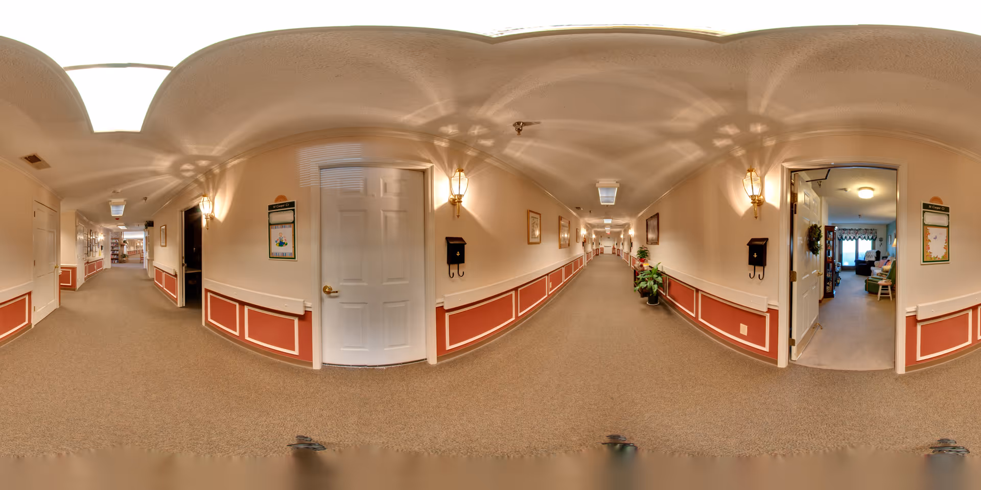 A long, well-lit hallway in a senior living facility with beige walls and carpeted floors. The walls have red and white wainscoting and are decorated with framed pictures and wall sconces. Several doors line the hallway, one of which is open, revealing a cozy room with furniture and decorations. There are plants placed along the hallway, and a bulletin board is visible on the wall.