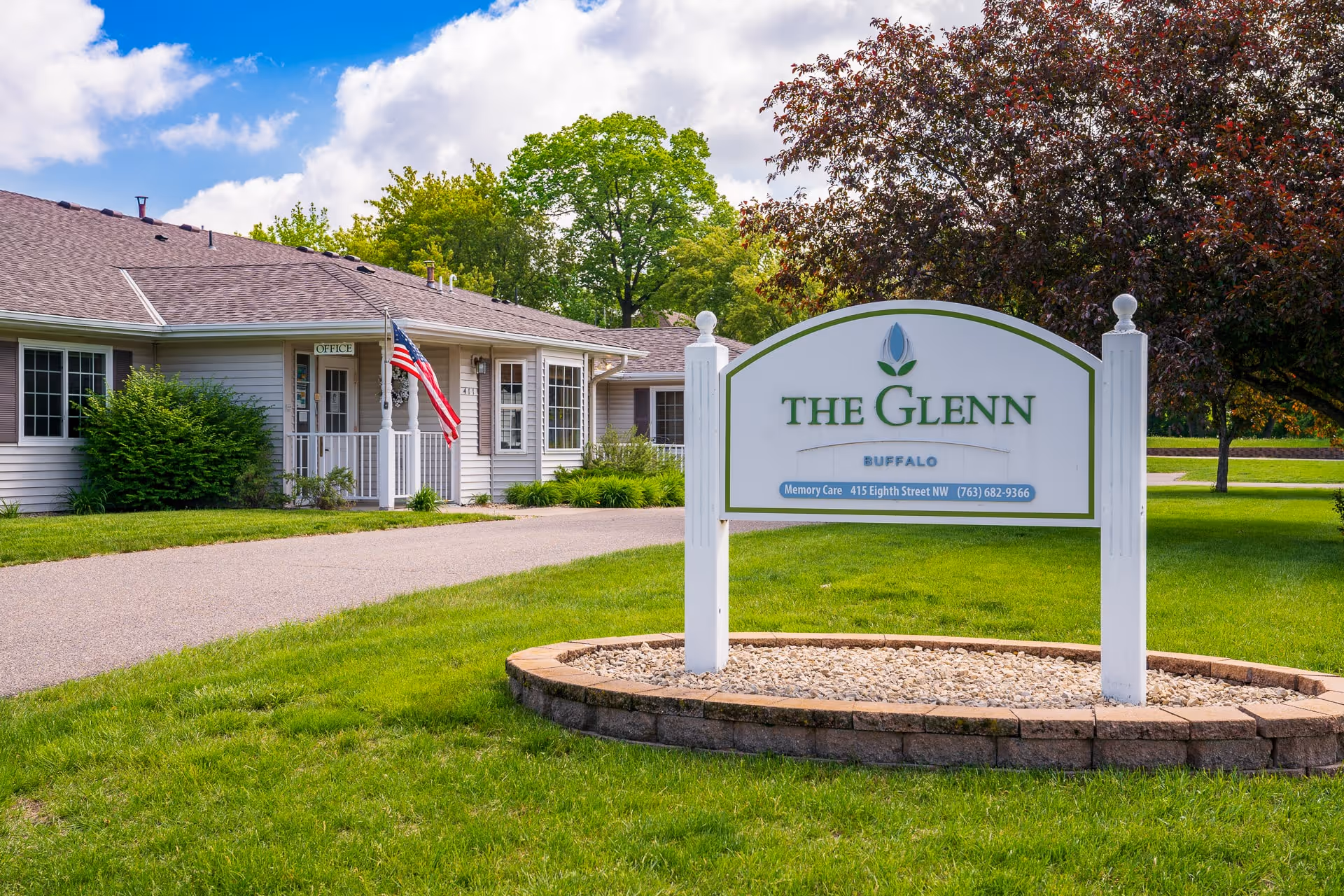 Exterior view of The Glenn Buffalo facility with a white sign displaying the facility name and contact information. The building has beige siding, several windows, an American flag near the entrance marked 'Office', and is surrounded by green grass and trees under a partly cloudy sky.