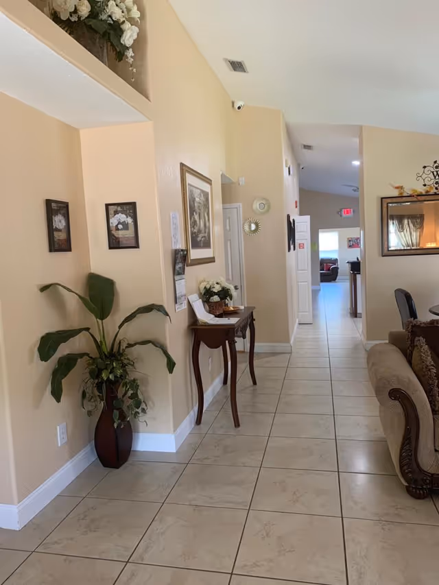 A hallway in a senior living facility with beige walls and tiled floor. On the left side, there is a tall plant in a dark vase and a small wooden table with flowers and papers on it. The walls are decorated with framed pictures and mirrors. On the right side, part of a brown upholstered sofa is visible. The hallway leads to another room with more seating visible in the distance.
