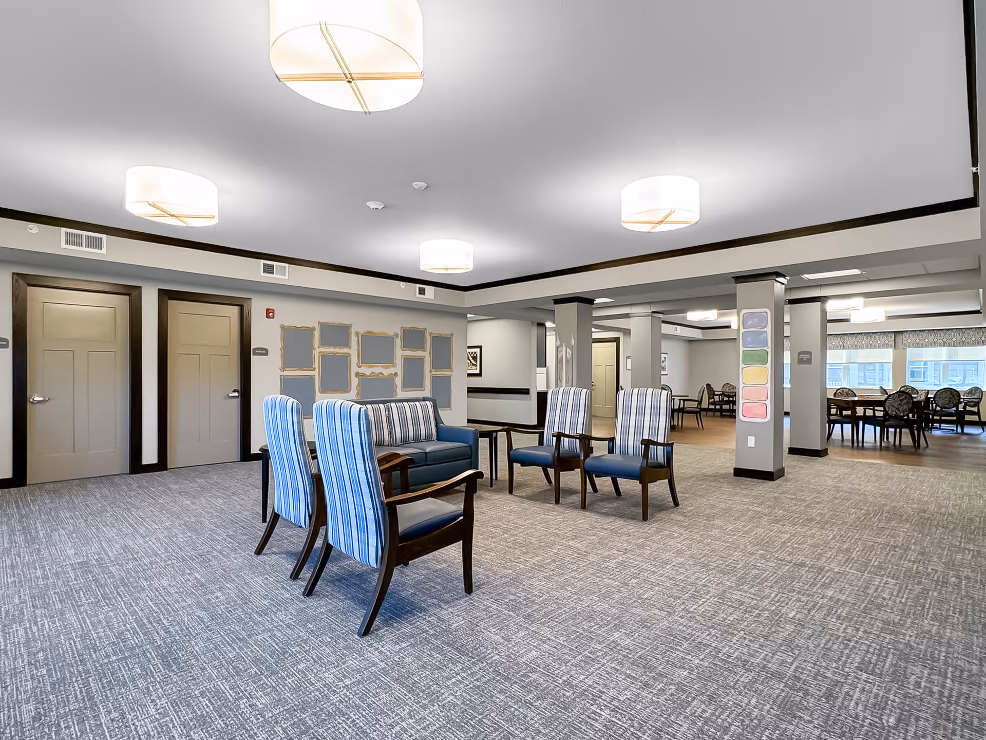 A spacious senior living facility common area with modern lighting fixtures on the ceiling. The room features a seating arrangement with four blue striped armchairs and a blue sofa with striped cushions. The walls are light-colored with dark trim, and there are multiple empty picture frames on one wall. In the background, there are additional tables and chairs near large windows allowing natural light to enter.