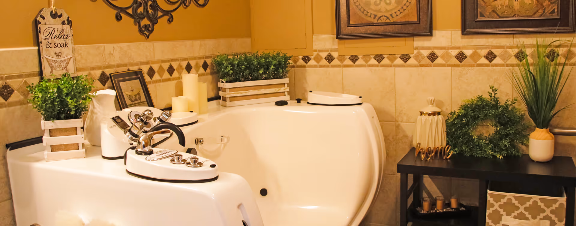 A cozy bathroom featuring a white walk-in bathtub with chrome fixtures, surrounded by decorative plants, candles, and framed artwork on beige tiled walls with a diamond pattern border. A small black table holds additional greenery and decorative items.