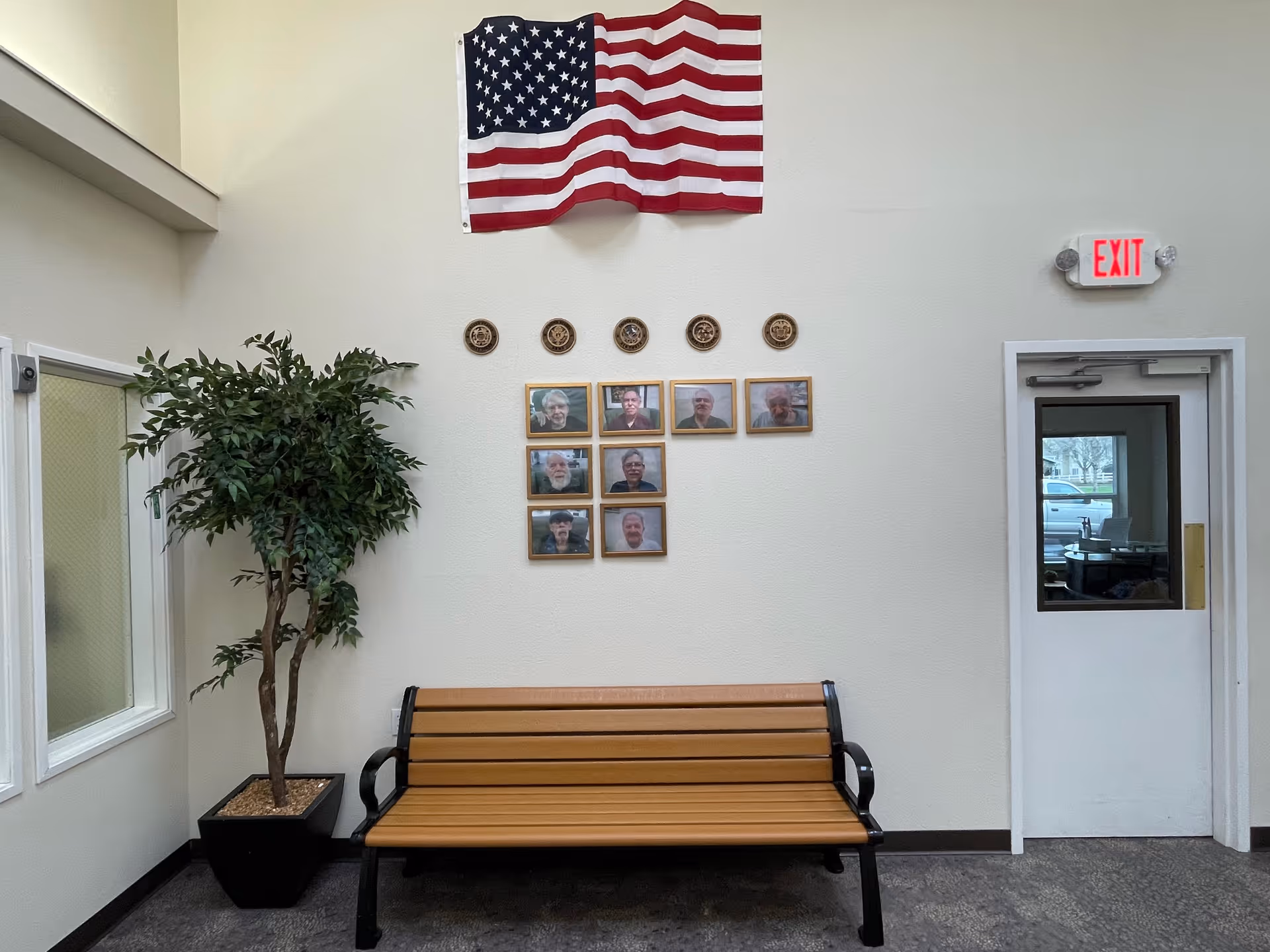 Interior wall of a senior living facility with an American flag hanging above a row of framed portraits and circular plaques. Below the portraits is a wooden bench with black metal armrests. To the left of the bench is a potted artificial tree, and to the right is a white door with a window and an illuminated red EXIT sign above it.