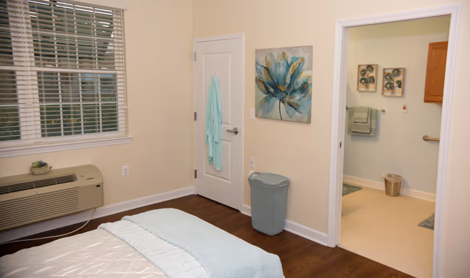 Light, neatly furnished bedroom with a bed, window, wall art, laundry basket, and an open door leading to a bathroom.