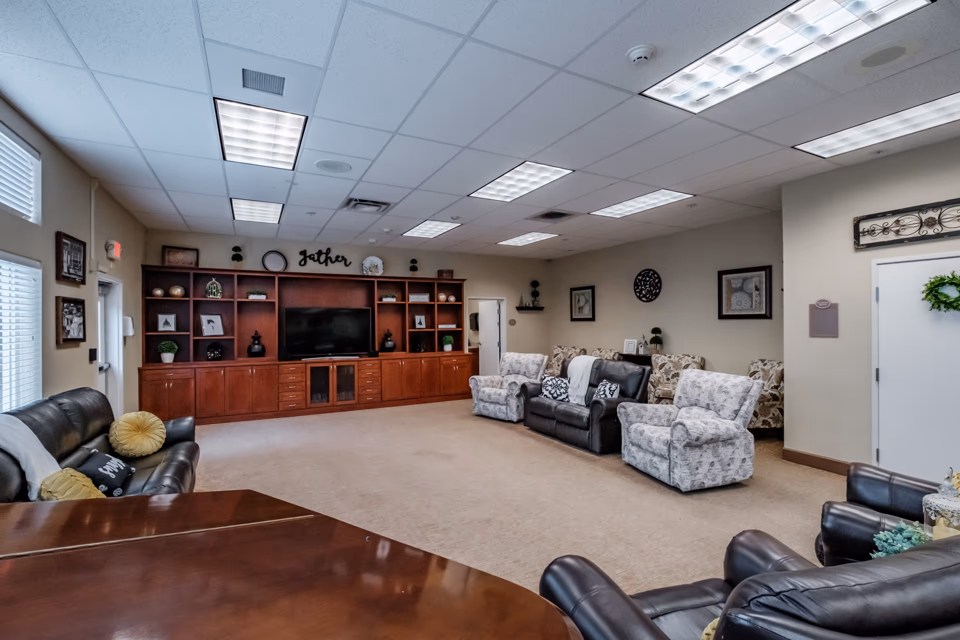 A spacious living room area in a senior living facility with multiple comfortable chairs and sofas arranged around a large wooden entertainment center with a flat-screen TV. The room has beige walls, carpeted floor, and ceiling lights. Decorative items and framed pictures adorn the walls and shelves.