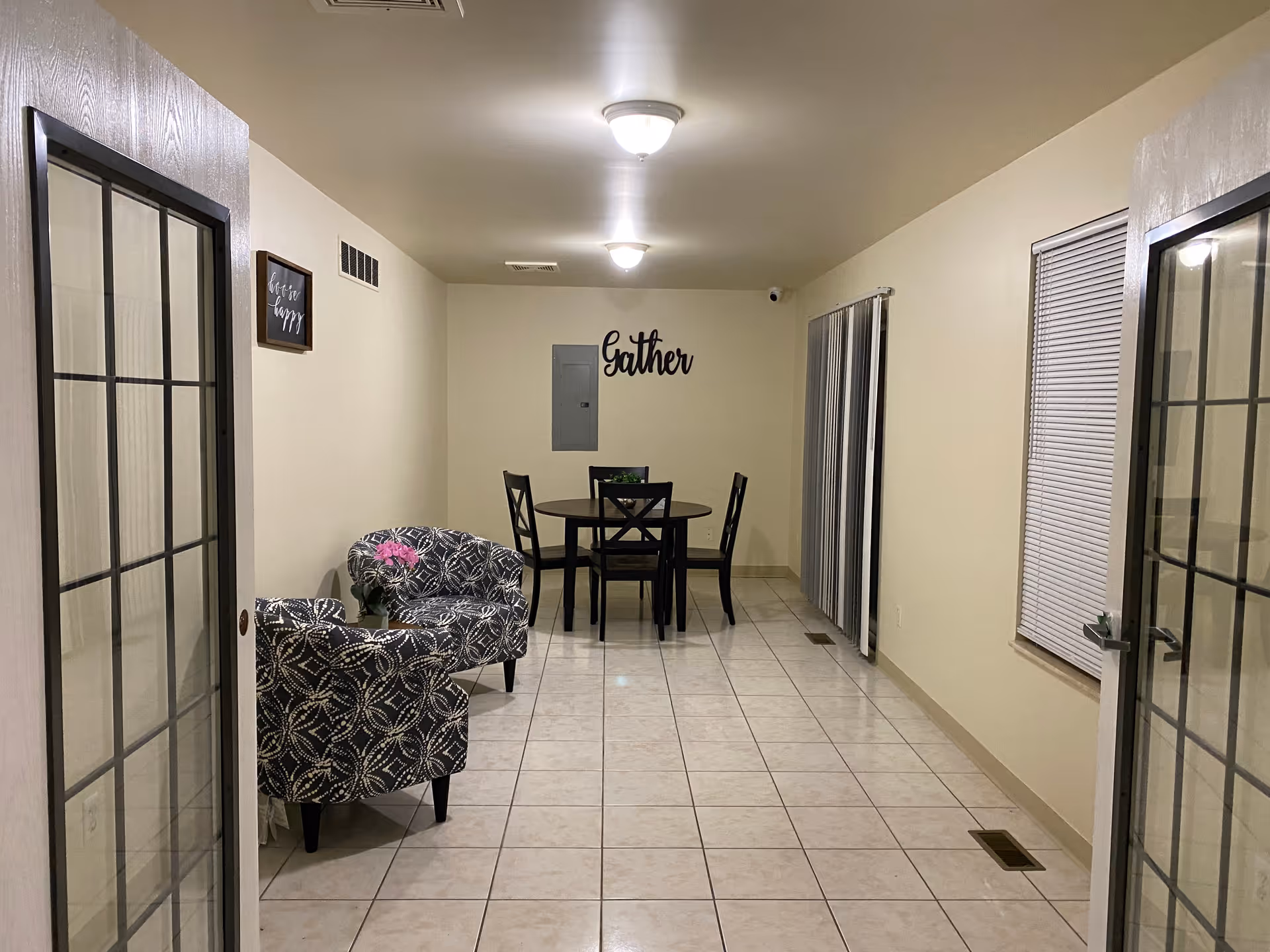 Interior view of a room with tiled floor, two patterned armchairs with a small table and flowers between them on the left, and a round dining table with four chairs at the far end. The walls are light-colored with a wall decoration that says 'Gather' above an electrical panel. There are two ceiling lights and windows with blinds and vertical blinds on the right side.