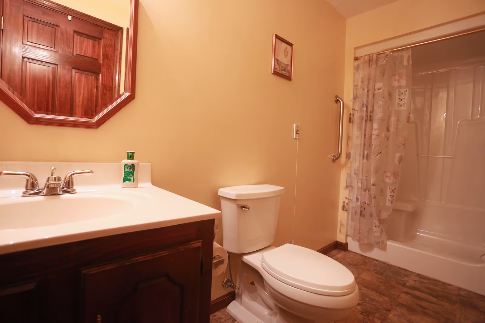 A bathroom with a white toilet, a sink with a silver faucet, a wooden framed mirror above the sink, a shower with a curtain featuring a butterfly and floral pattern, a grab bar on the wall next to the shower, and a small framed picture on the wall.