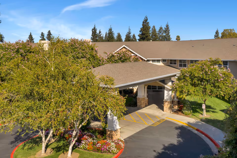 Entrance area of Dale Commons senior living facility with a covered drop-off zone, surrounded by green trees and colorful flower beds under a clear blue sky.