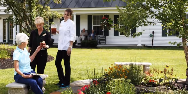 Three women socialize in a landscaped courtyard in front of a white senior living building, with one seated on a bench and two standing near flowerbeds.