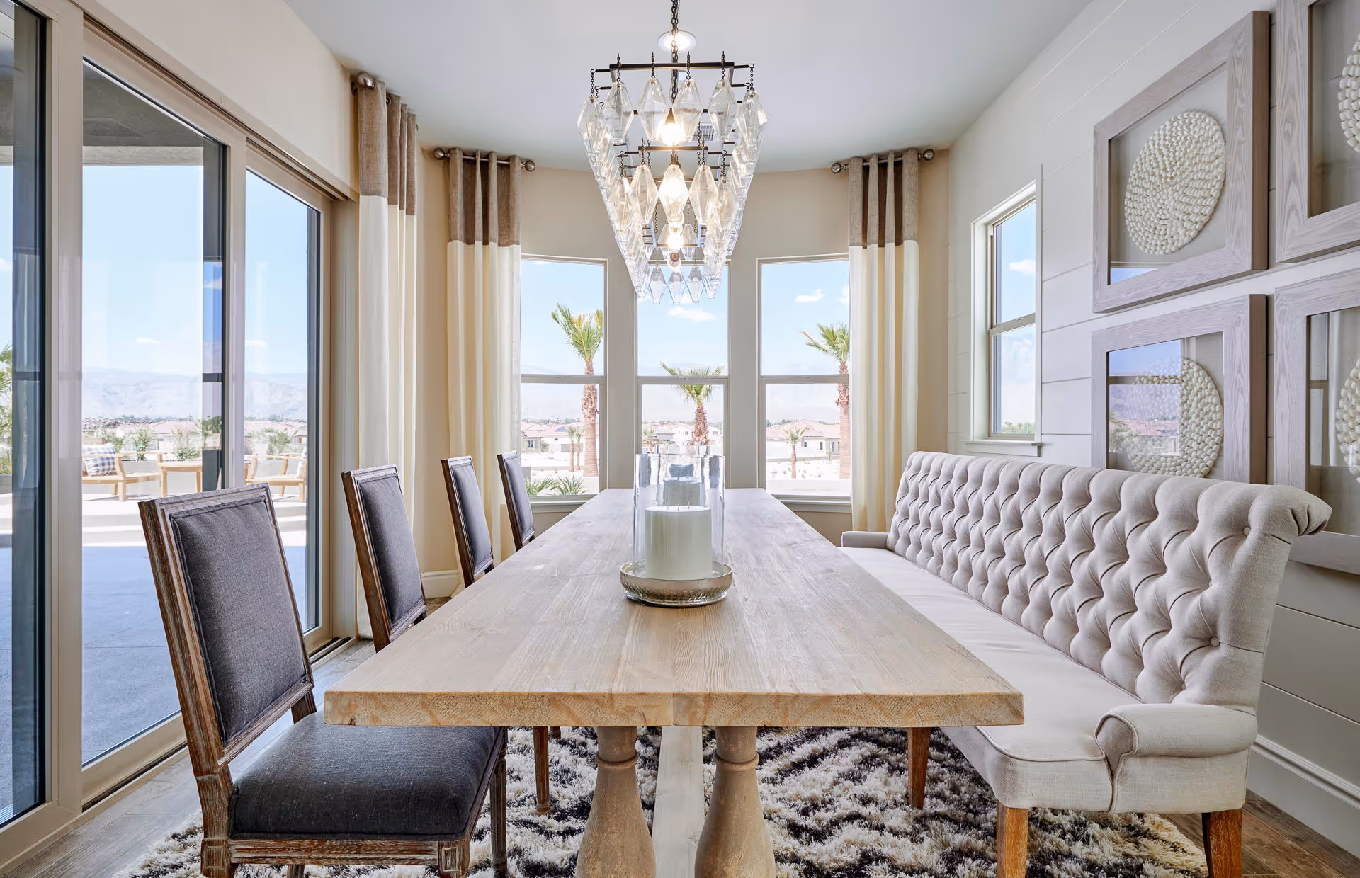 Bright dining room with a long wooden table surrounded by four dark cushioned chairs on one side and a tufted beige bench on the other. Large windows with beige and brown curtains let in natural light and offer a view of palm trees and distant mountains. A modern chandelier hangs above the table, and decorative wall art is displayed on the right wall. Sliding glass doors open to an outdoor patio area.