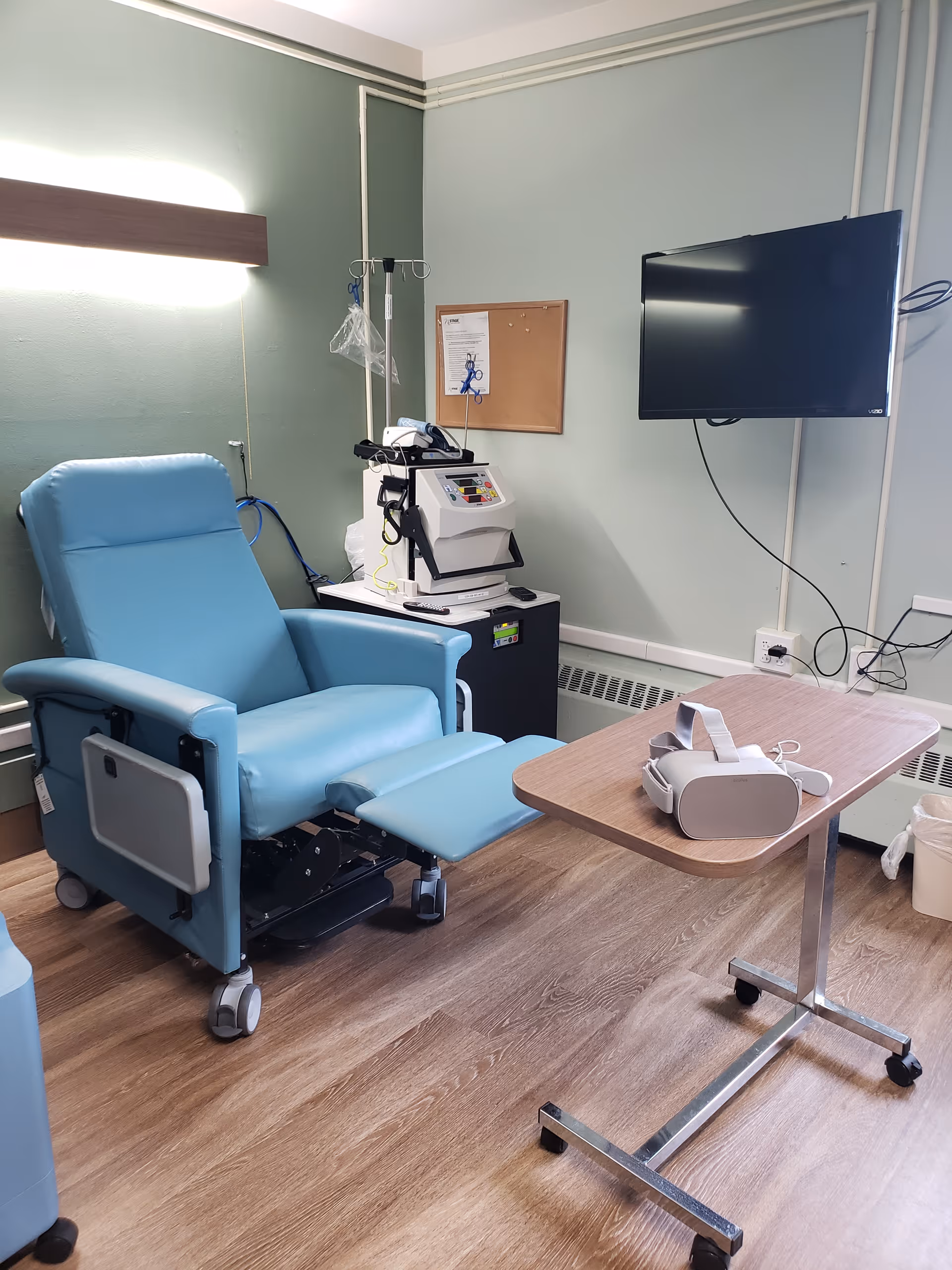 A medical treatment room with a light blue recliner chair, an IV pole, a dialysis machine, a wall-mounted flat screen TV, a cork bulletin board, and a small overbed table with a virtual reality headset on it. The room has light green walls and wood-patterned flooring.