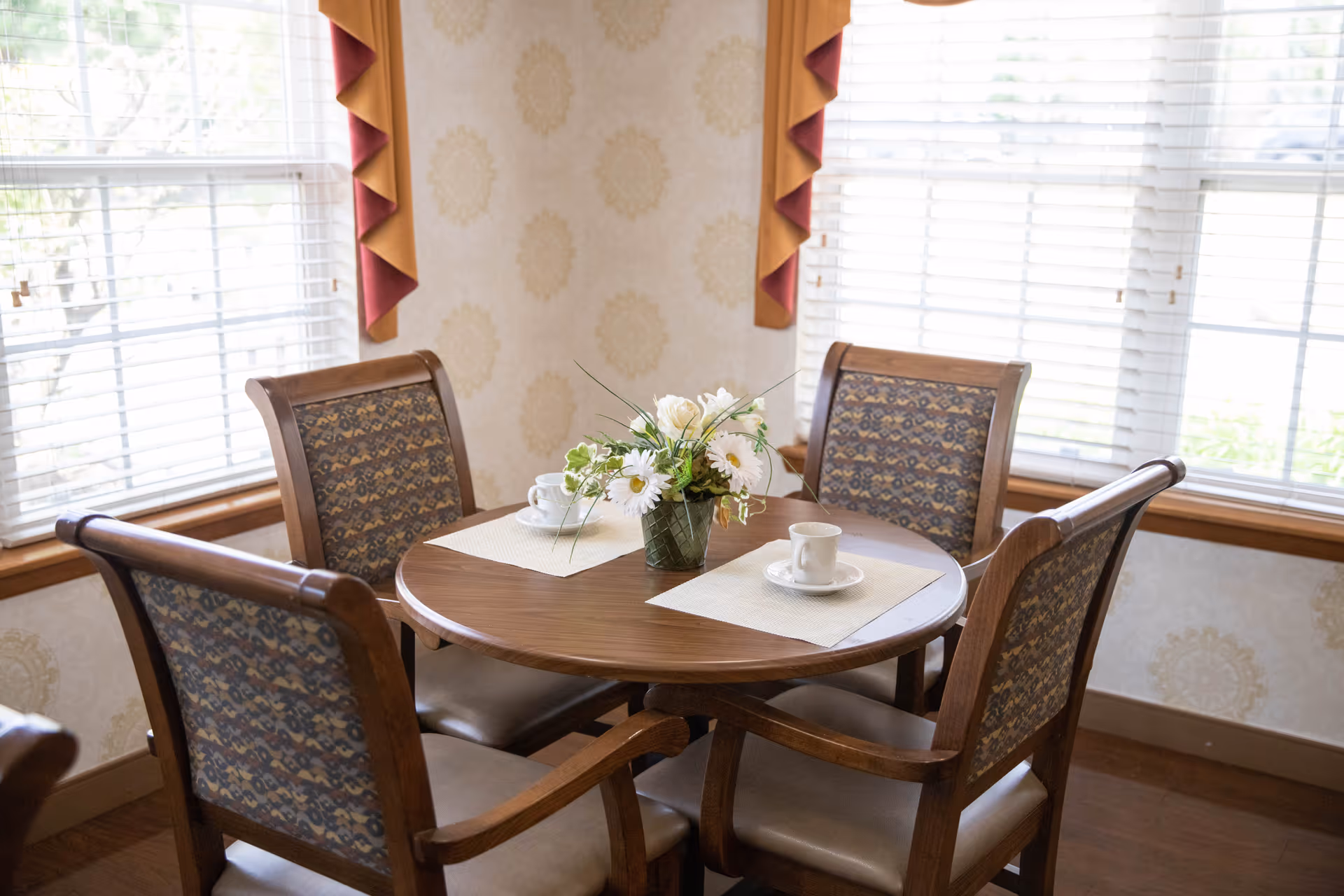 A round wooden dining table set with four upholstered chairs in a corner with two large windows covered by white blinds and decorative valances. The table has two white cups and saucers and a small floral arrangement in the center.