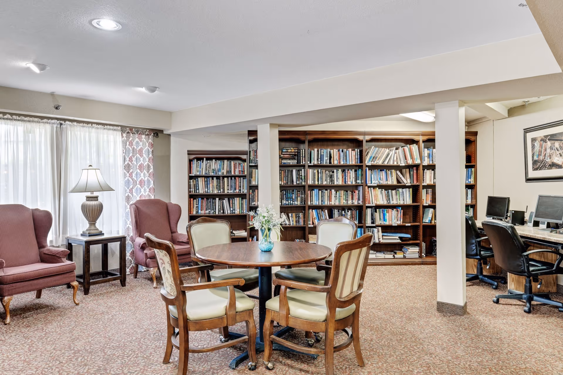 A cozy common area with a round wooden table surrounded by four cushioned chairs, two upholstered armchairs with a side table and lamp between them, large bookshelves filled with books, and a computer workstation with three chairs along the wall.