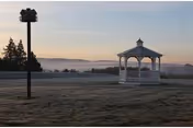 A white gazebo on a frosty lawn at sunrise with a birdhouse on a post and misty hills in the background.