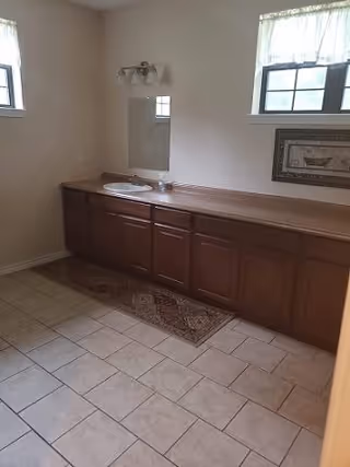 A bathroom with a long wooden vanity featuring a single sink and a mirror above it. The room has tiled flooring, two small windows with white curtains, and a framed picture on the wall.