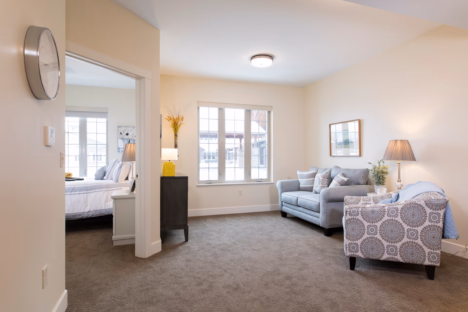 A bright and cozy living room with a gray sofa and a patterned armchair, both adorned with cushions. A floor lamp with a beige shade stands next to the armchair. A framed picture hangs on the wall above the sofa. The room has a large window letting in natural light. To the left, an open doorway leads to a bedroom with a neatly made bed, bedside table, and lamp. A clock is mounted on the wall near the doorway.