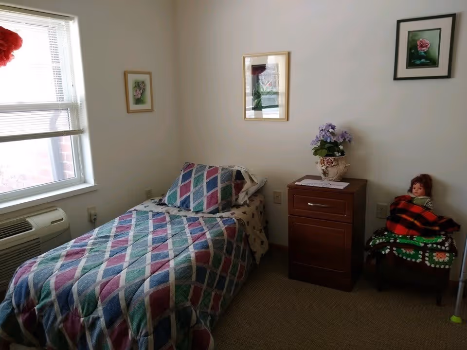 A small bedroom with a single bed covered in a colorful diamond-patterned quilt and matching pillow. Next to the bed is a wooden nightstand with a decorative flower pot on top. On the right side, there is a chair with a doll wrapped in a red and black plaid blanket sitting on it. The walls are white and decorated with three framed floral pictures. A window with blinds is on the left side, and an air conditioning unit is installed below the window.