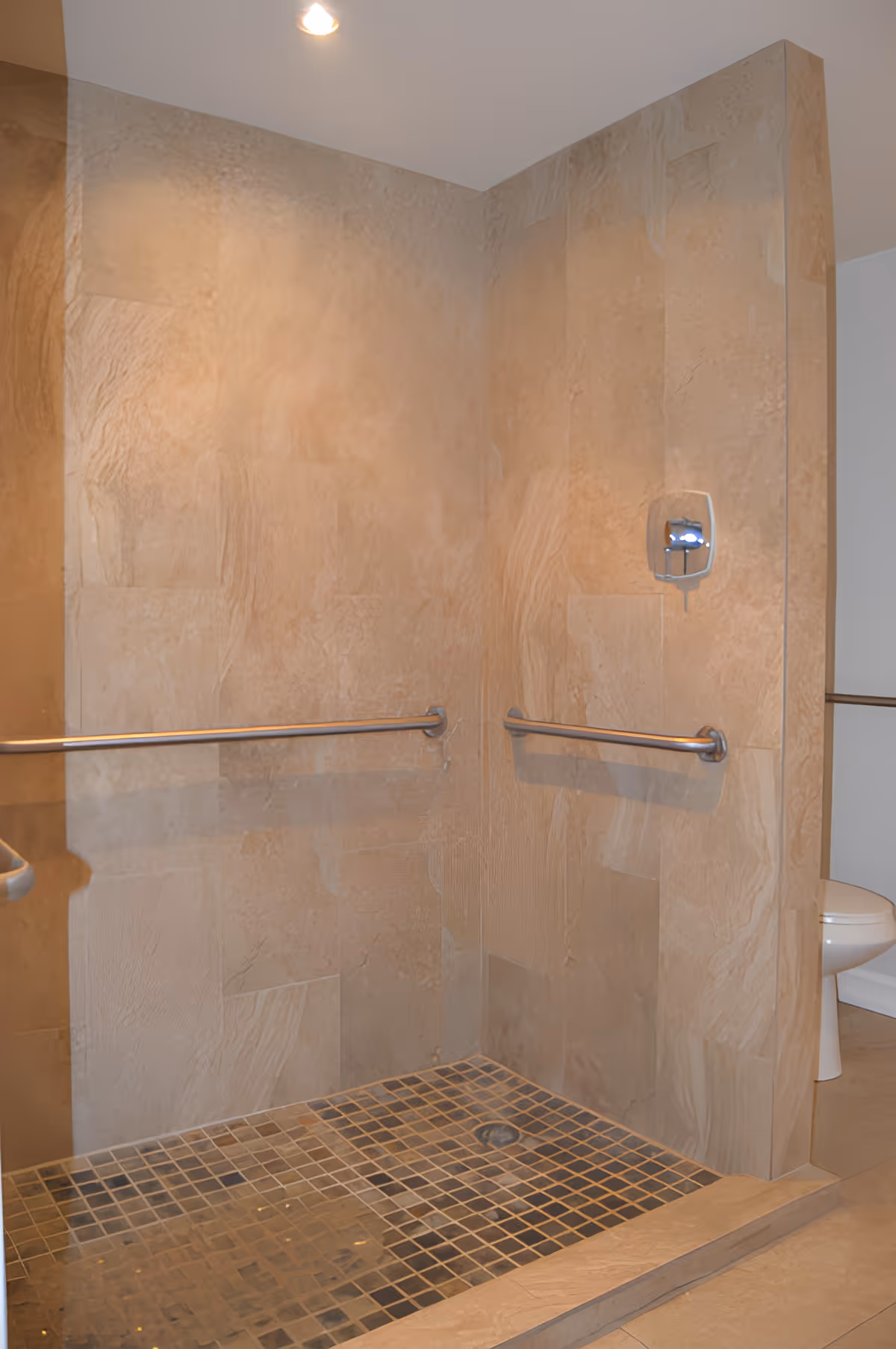 A walk-in shower with beige tiled walls and floor, equipped with two horizontal metal grab bars and a wall-mounted soap dispenser. A toilet is partially visible behind the shower wall.