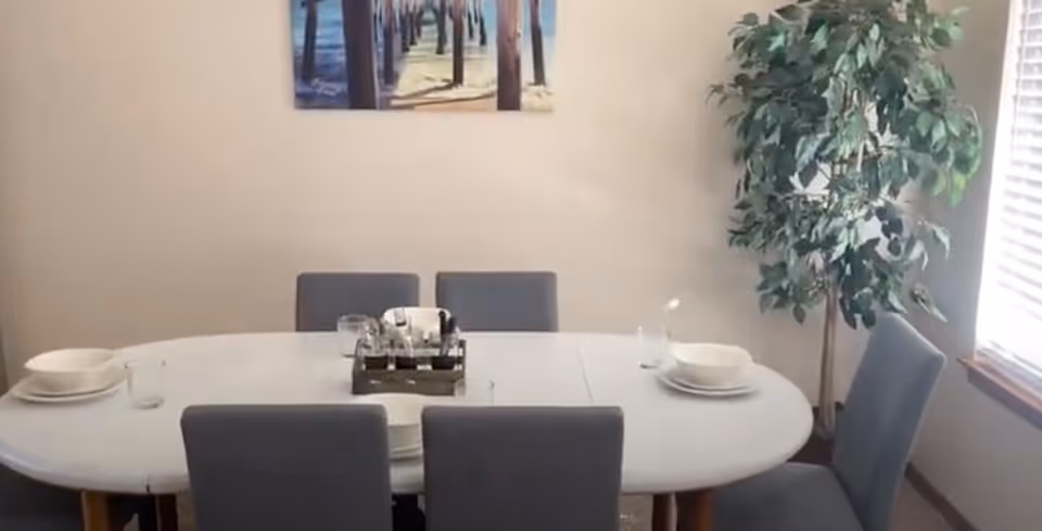 Oval dining table set with plates and glasses surrounded by gray chairs, a large potted plant, and wall art beside a sunlit window.