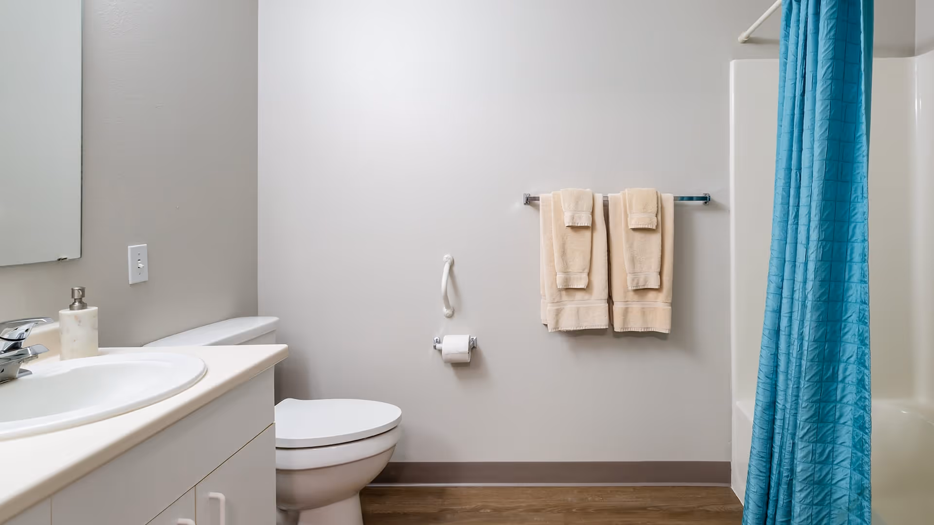 A clean bathroom featuring a white sink with a silver faucet and soap dispenser on the left, a white toilet in the center, beige towels hanging on a towel rack above a toilet paper holder, and a bathtub with a bright blue shower curtain on the right.
