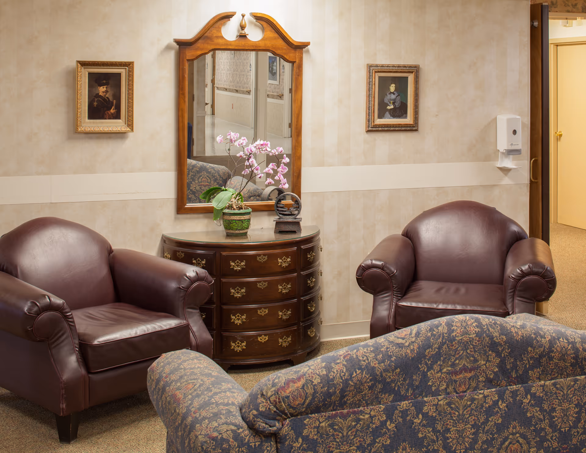 A cozy sitting area with two dark brown leather armchairs and a patterned fabric sofa. Between the armchairs is a wooden chest of drawers with brass handles, topped with a potted orchid and a decorative item. Above the chest is a wooden-framed mirror reflecting a hallway. Two framed portraits hang on the beige wallpapered walls, and a wall-mounted soap dispenser is visible near an open doorway.