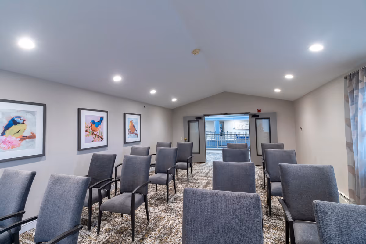 A small meeting or conference room with rows of gray upholstered chairs arranged facing forward. The room has a carpeted floor with a patterned design, light gray walls, and a vaulted ceiling with recessed lighting. Three framed pictures of colorful birds are hung on the left wall. Double doors at the far end of the room open to a hallway with railings and additional artwork visible.