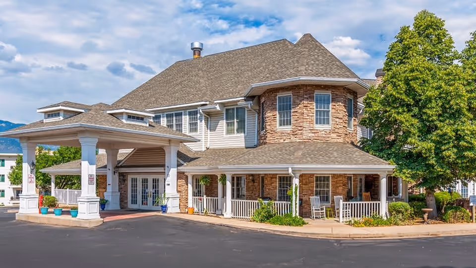 Exterior view of a senior living facility building with a covered entrance, stone and siding facade, multiple windows, and a porch with rocking chairs. There are potted plants near the entrance and a large tree on the right side under a partly cloudy sky.