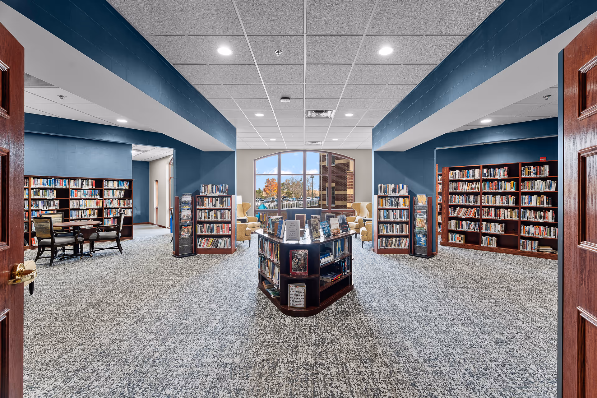 Interior view of a library room with blue walls and carpeted floor. There are multiple wooden bookshelves filled with books, a central display bookshelf, and a seating area with yellow armchairs near a large window showing an outdoor view.