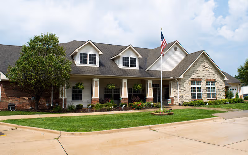 Front exterior of a single-story senior living building with a covered porch, American flag, and landscaped lawn.